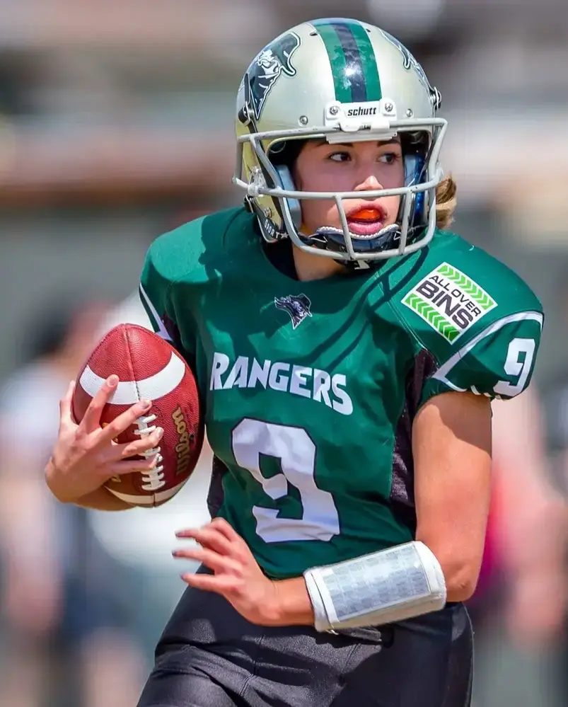 A high-intensity action shot of a female American football player wearing a dark green "RANGERS" jersey with the number 9, sprinting with the ball while wearing a silver helmet and protective mouthguard.