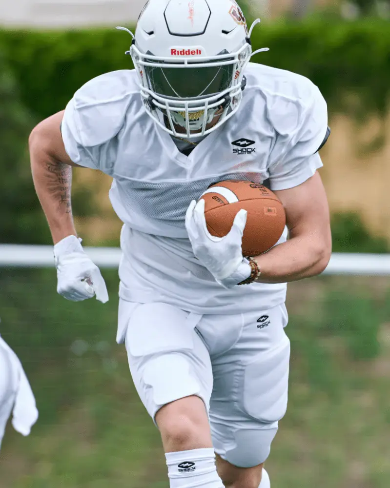 A high-action shot of an American football player in a clean white practice jersey and padded pants, sprinting while holding the football, wearing a red Riddell helmet and white gloves.