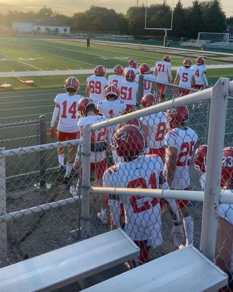 A group of American football players in full uniform white jerseys with large red numbers and logos, white pants, red helmets, and red socks walks down a set of stadium stairs and through a chain-link fence gate onto a green grass field during a sunset game.