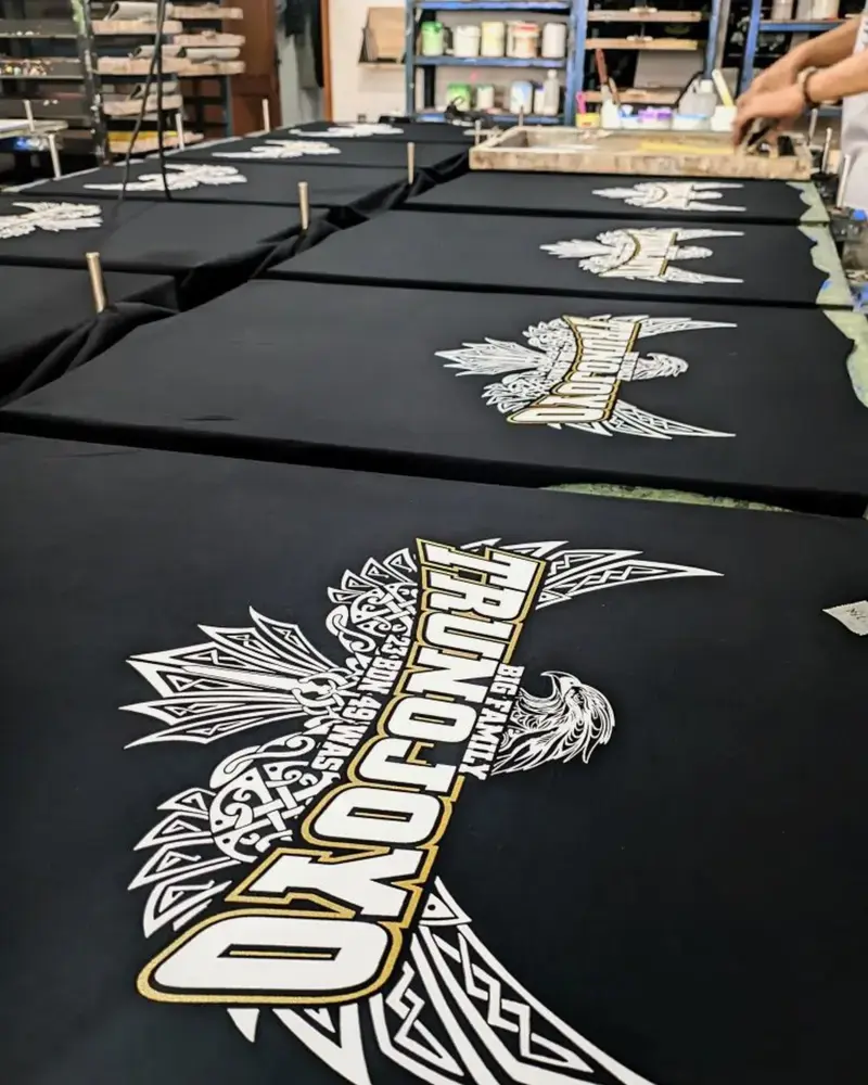 Multiple black t-shirts laid out on a printing press table, each featuring a large white and gold eagle graphic with bold text being printed in a workshop setting.