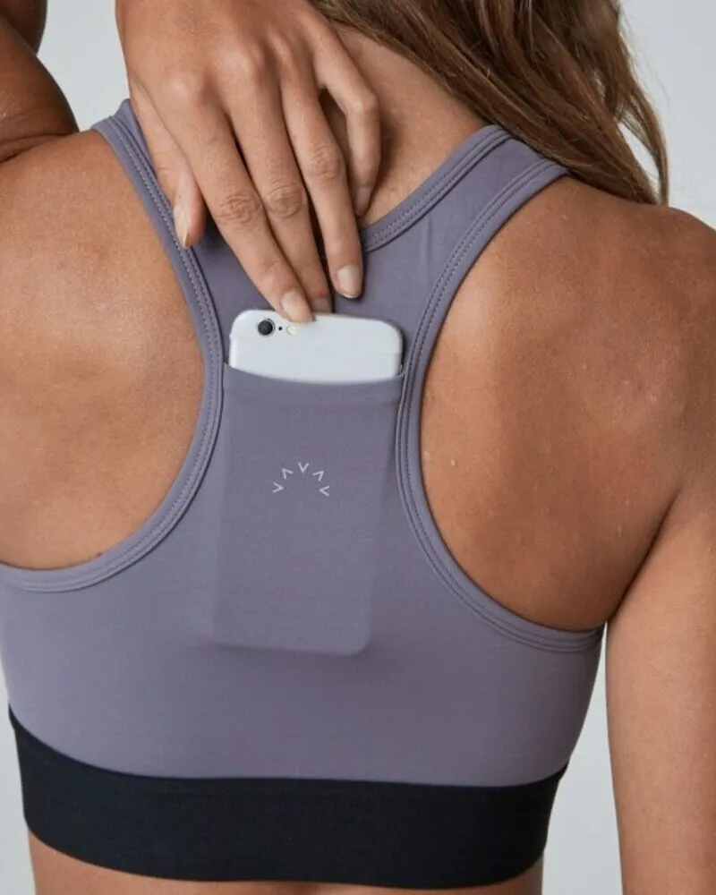 A back view of a person wearing a purple racerback sports bra with a black elastic band, featuring a functional pocket on the racerback panel with a smartphone being tucked inside.