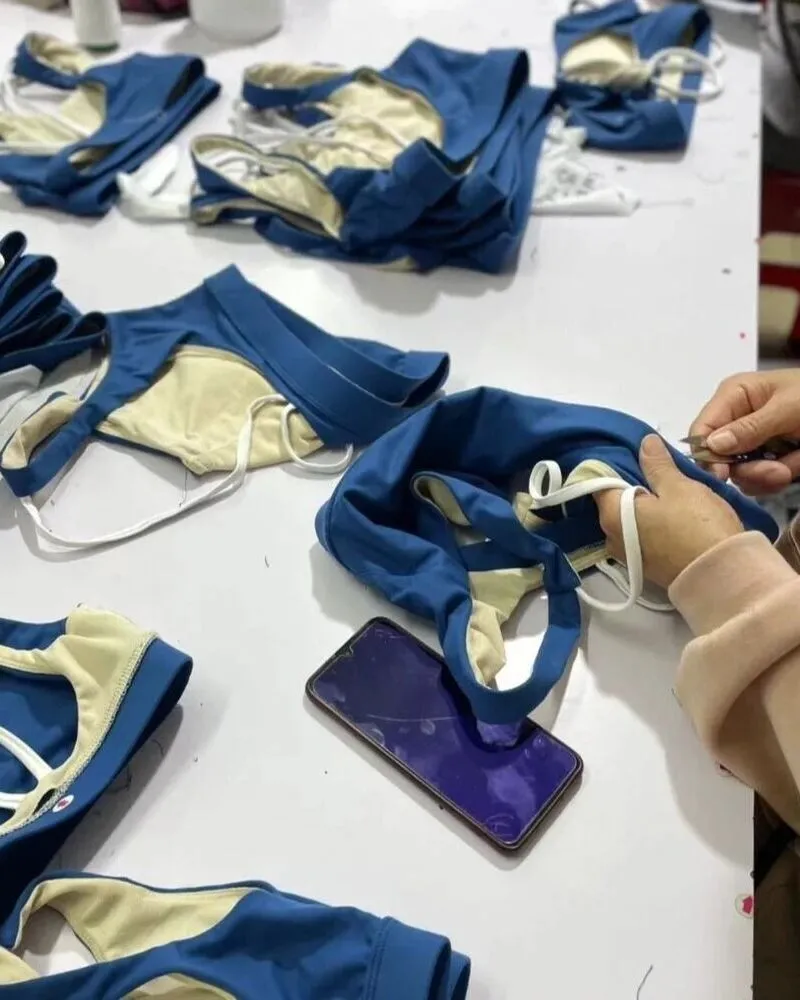 A close-up view of several blue sports bras with white elastic straps being finished on a white habit fit sports factory table, with a worker's hands using small scissors to trim threads.