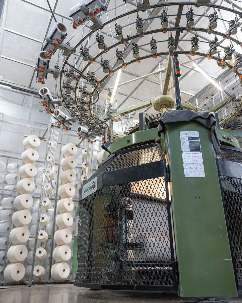 A large-scale circular industrial knitting machine in a textile factory, surrounded by tall racks of white yarn spools ready for fabric production.